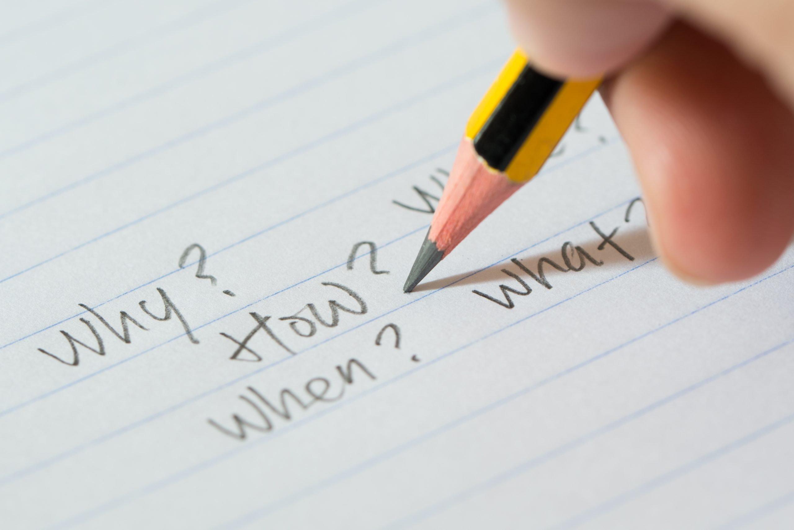 A person writing "Why?", "How?", and "When" in pencil on a piece of paper