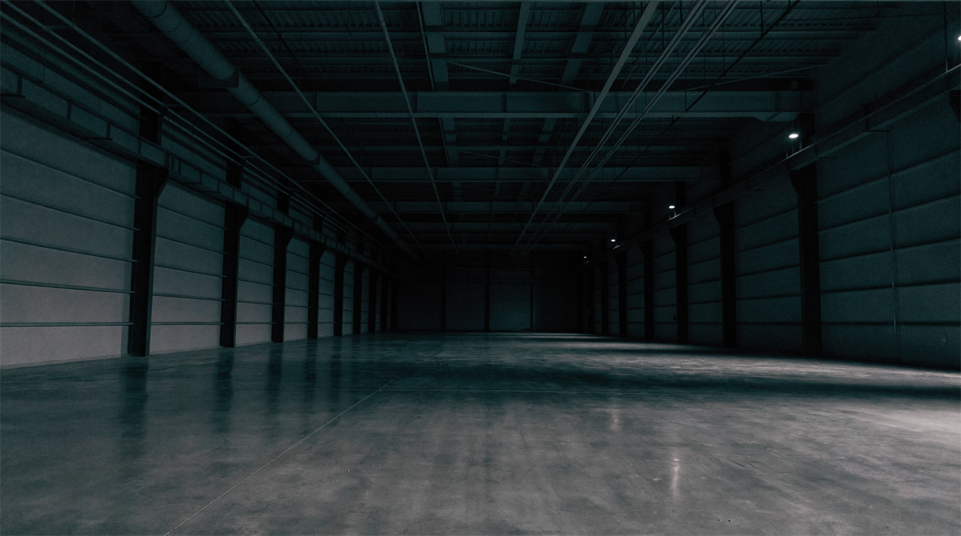 A long industrial corridor with polished concrete floor and steel structure receding into shadow, sparsely lit and largely empty