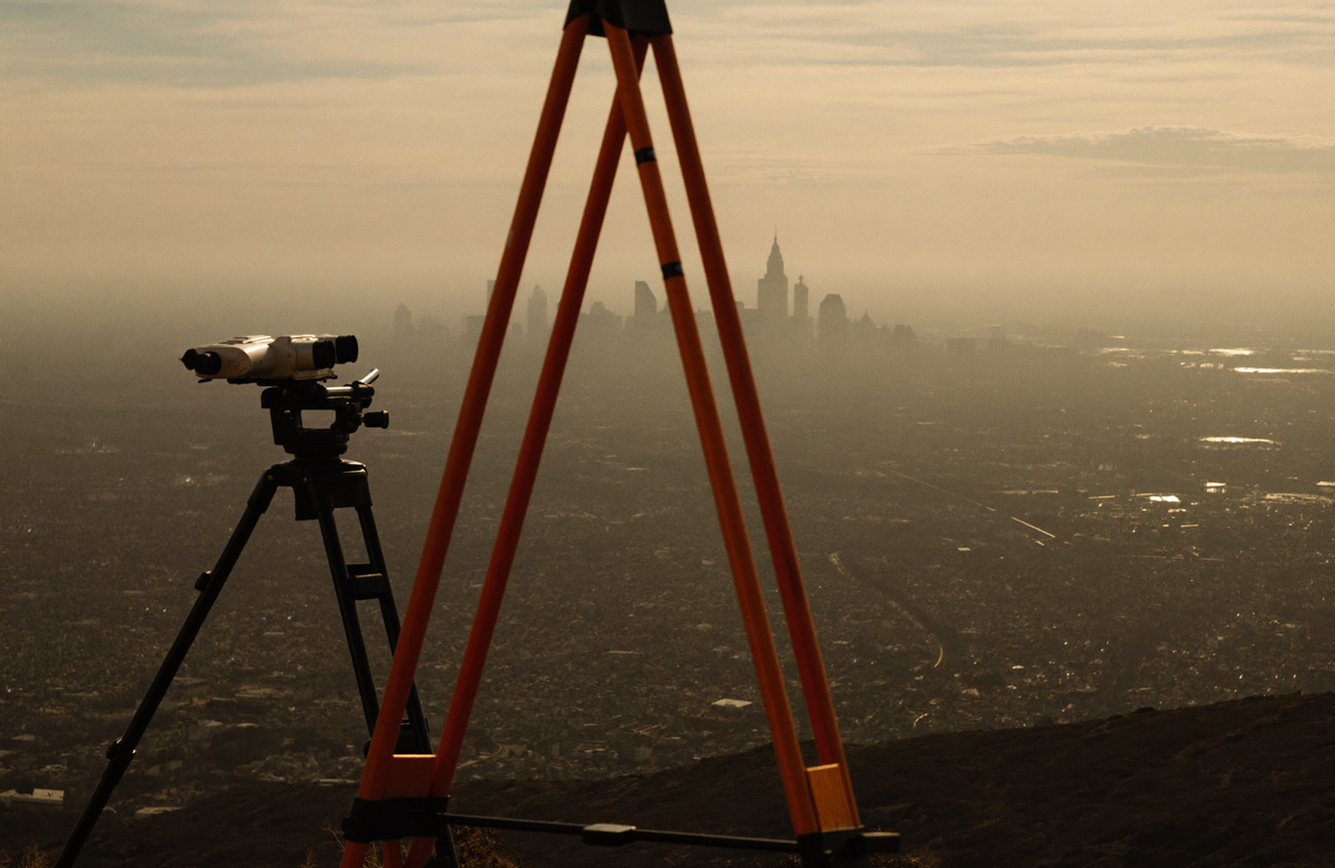 A surveyor’s theodolite on a tripod aimed at a distant hazy city skyline in golden morning light